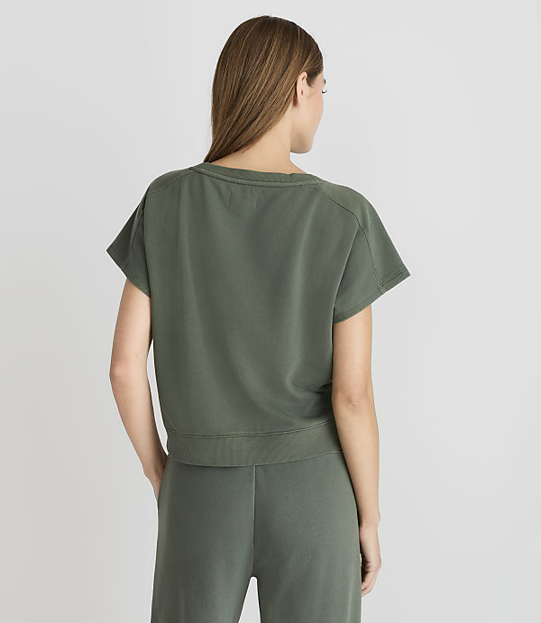 A woman in a green t-shirt and pants stands against a plain gray background.