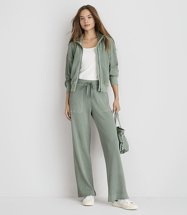 A woman in a sage green loungewear set stands against a plain white background, holding a matching bag.