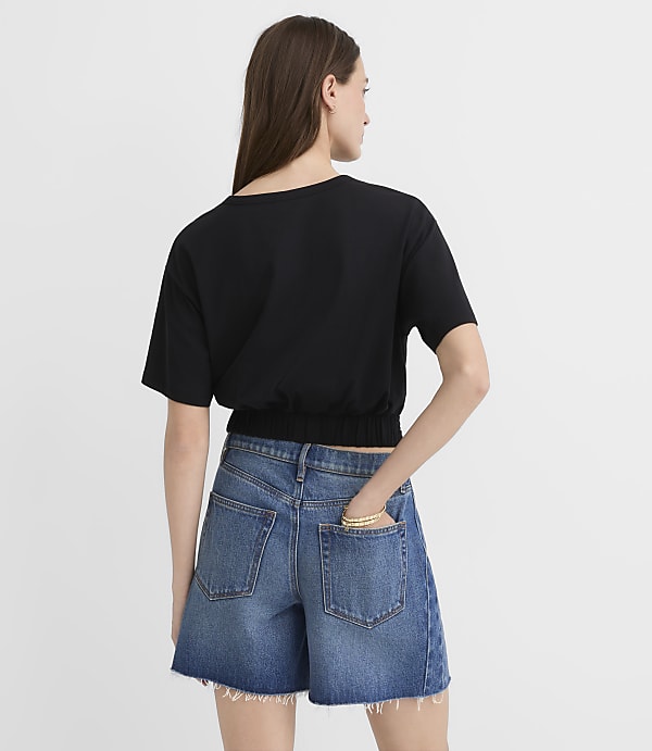 A woman in a black t-shirt and denim shorts stands against a plain white background.