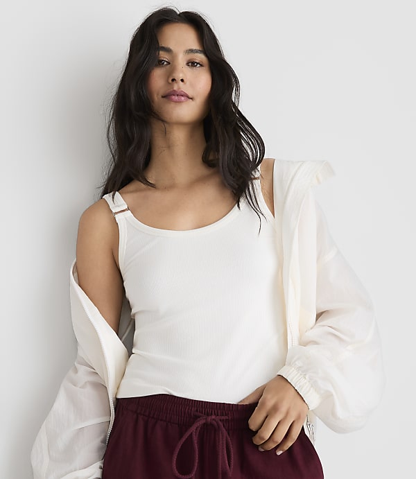 A woman in a white tank top and maroon pants stands against a plain white background.