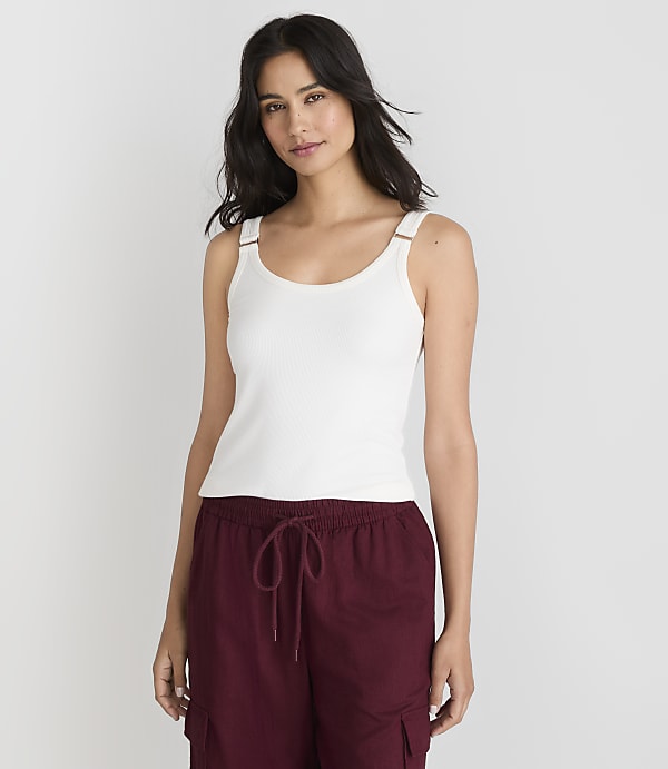 A woman in a white tank top and maroon pants stands against a plain white background.