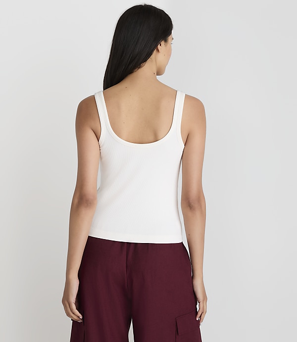 A woman in a white tank top and maroon pants stands against a plain white background.