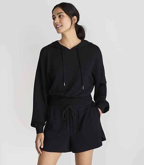 A woman in a black hoodie and shorts stands against a plain white background.