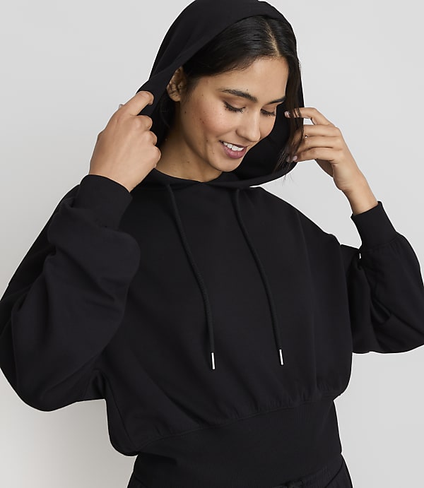 A smiling woman in a black hoodie adjusts the hood against a plain gray background.