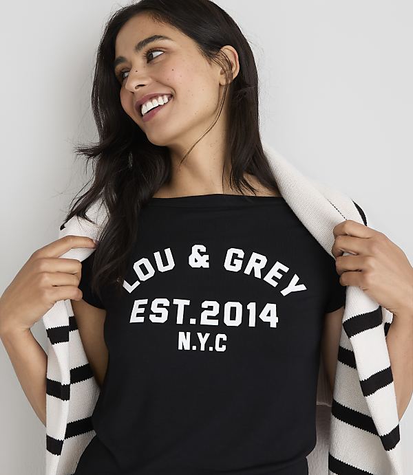 A smiling woman in a black t-shirt with "LOU & GREY EST. 2014 N.Y.C." printed on it, draped with a striped scarf, stands against a plain white background.