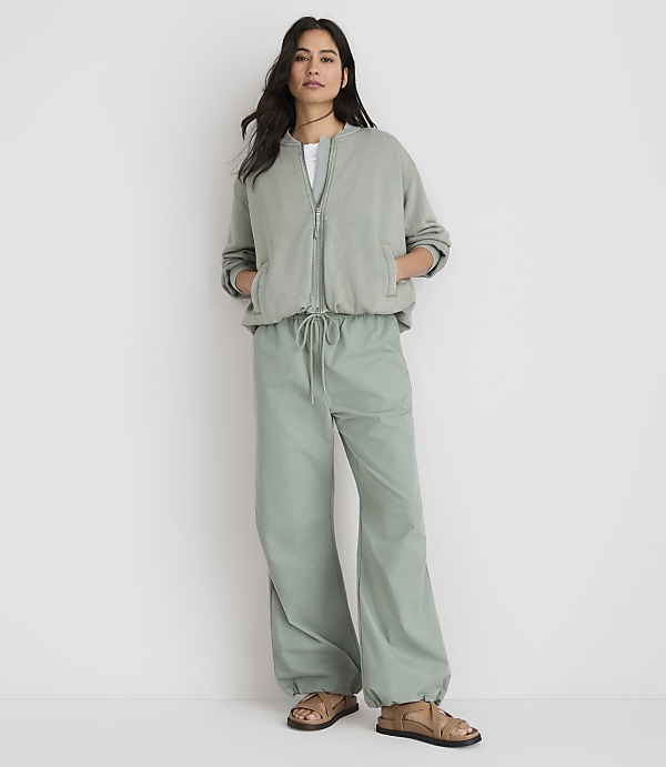 A woman stands in the foreground wearing a sage green bomber jacket and matching wide-leg pants, against a plain white background.