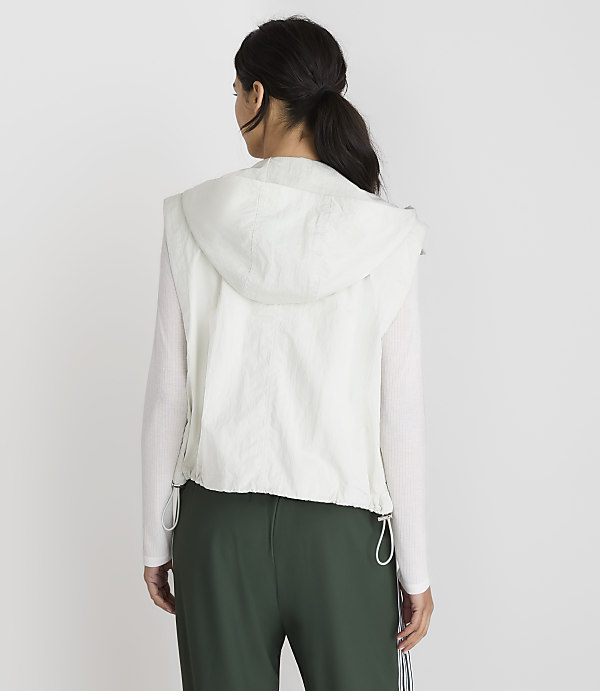 A woman in a white hooded vest and green pants stands against a plain white background.