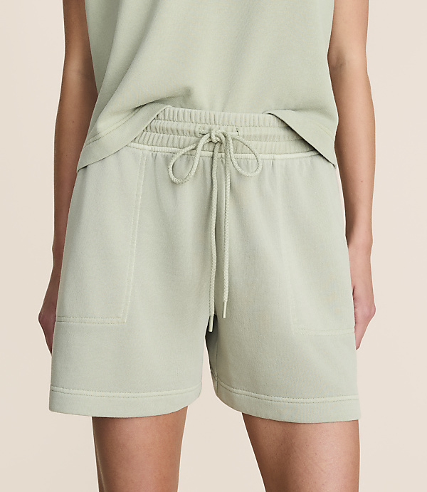 A person models light green sweat shorts with a drawstring waist and pockets in the foreground, against a plain, light pink background.