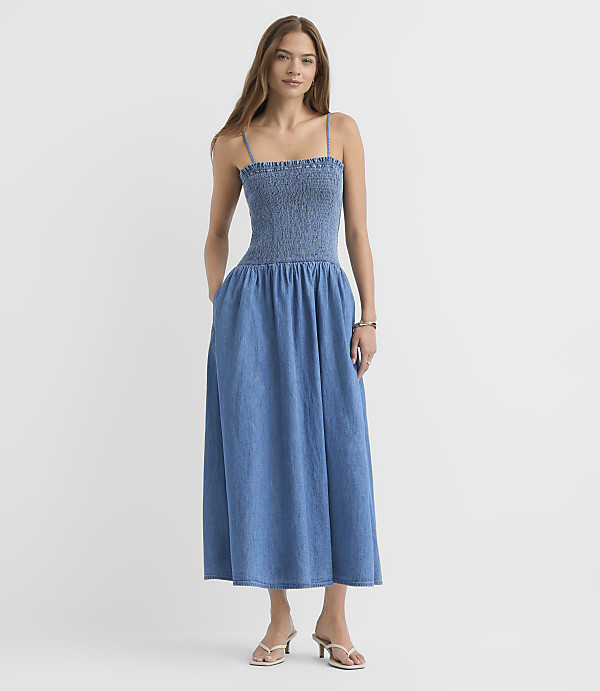 A woman in a blue denim midi dress with smocked bodice and pockets stands against a plain white background.