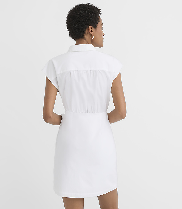 A woman in a white shirt dress stands against a plain white background.