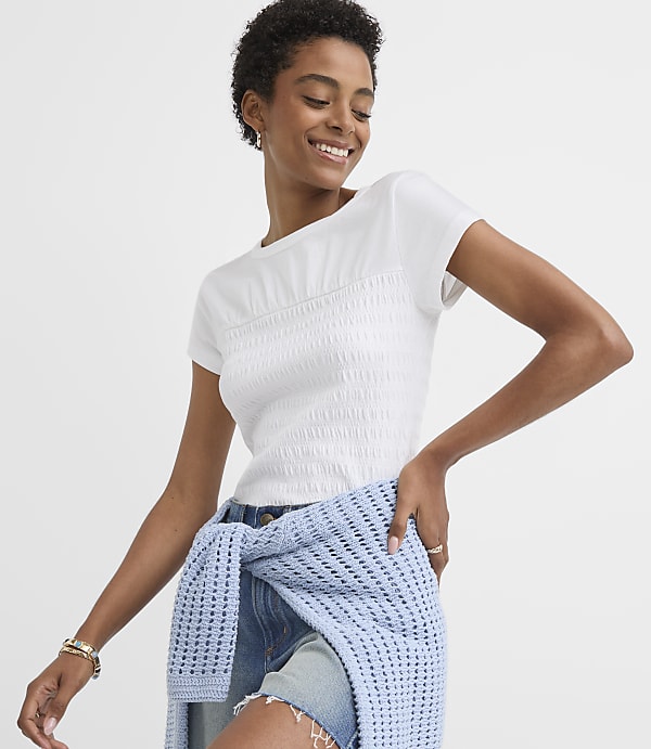 A smiling woman in a white smocked top and denim shorts is leaning against a light blue textured sweater against a plain white background.