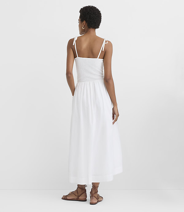 A woman in a white smocked sundress stands with her back to the viewer against a plain white background.