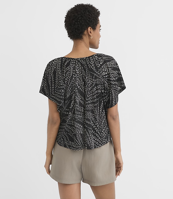A woman in a black patterned top and tan shorts stands against a plain white background.