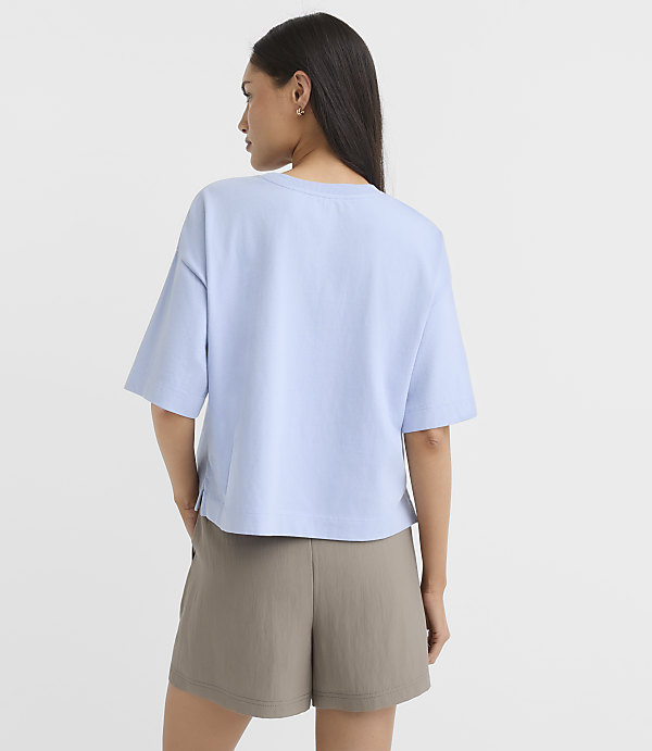 A woman in a light blue t-shirt and olive shorts stands against a plain white background.