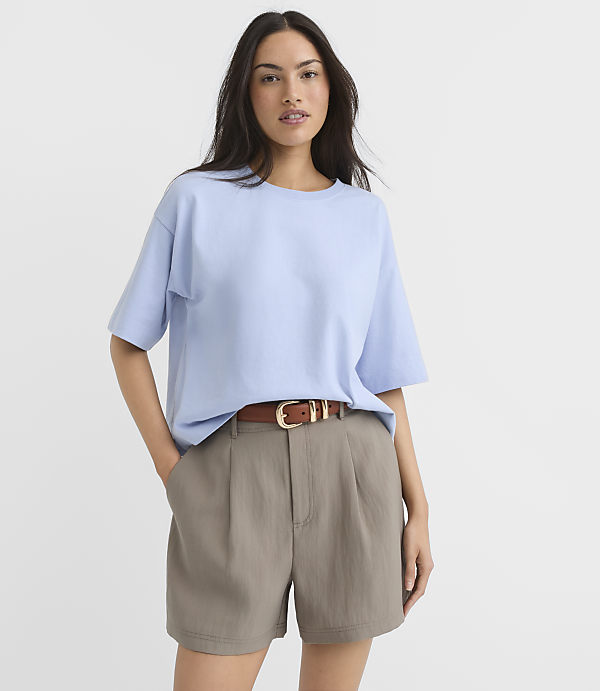 A woman in a light blue t-shirt and taupe shorts stands against a plain white background.
