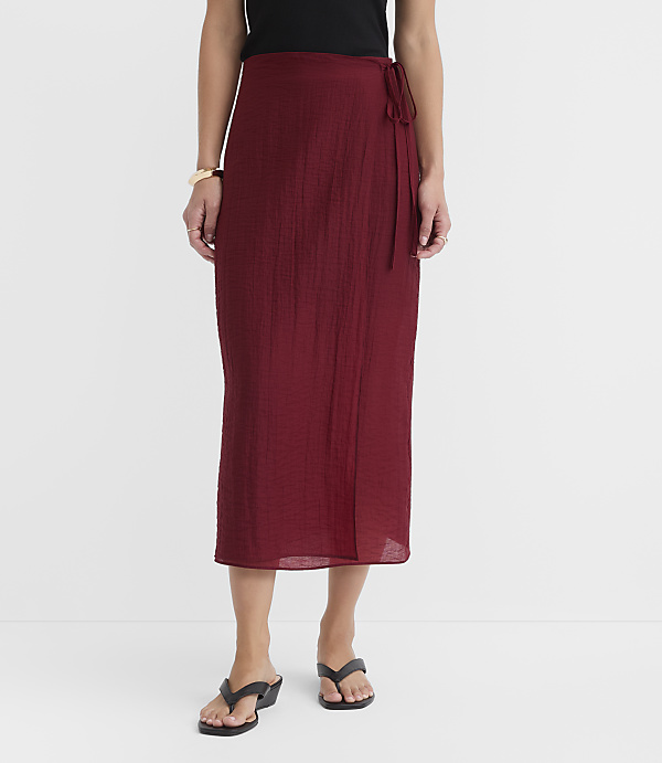 A woman in a maroon wrap skirt and black top stands against a plain white background.