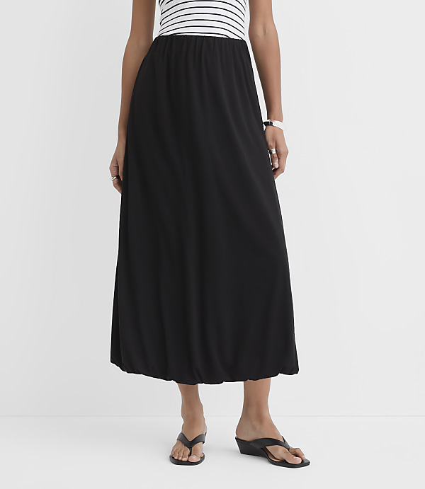 A woman in a black maxi skirt and a striped top stands against a plain white background.