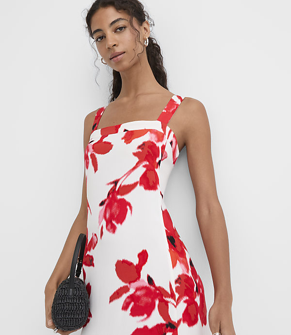A woman in a white floral dress with a black purse stands against a plain white background.