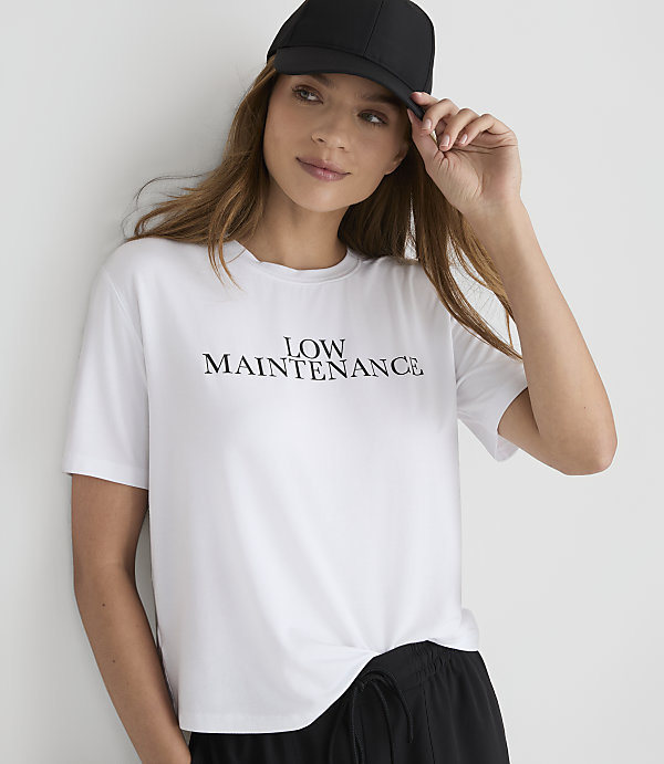 A woman in a white \"LOW MAINTENANCE\" t-shirt and black baseball cap poses against a plain white background.