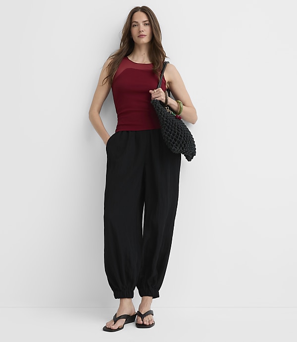 A woman in a maroon tank top and black pants stands against a plain white background, holding a black bag.