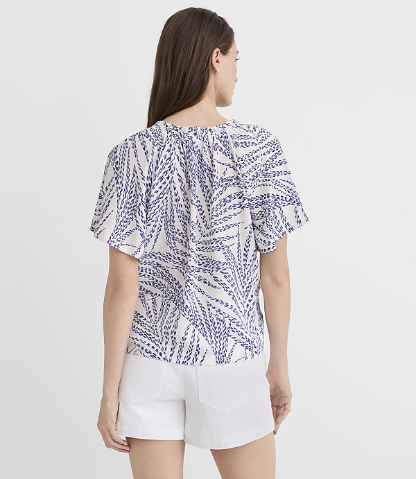 A woman in a white and blue patterned top and white shorts stands against a plain white background.