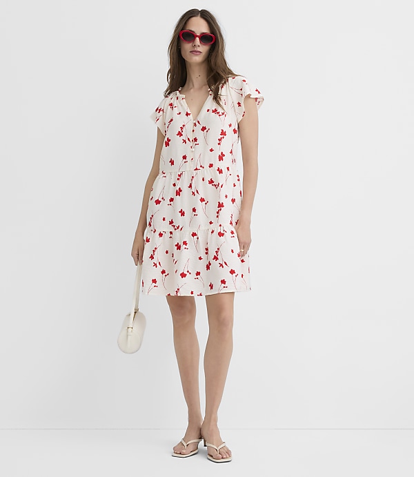 A woman in a white floral dress and red sunglasses stands against a plain white background, holding a cream-colored purse.