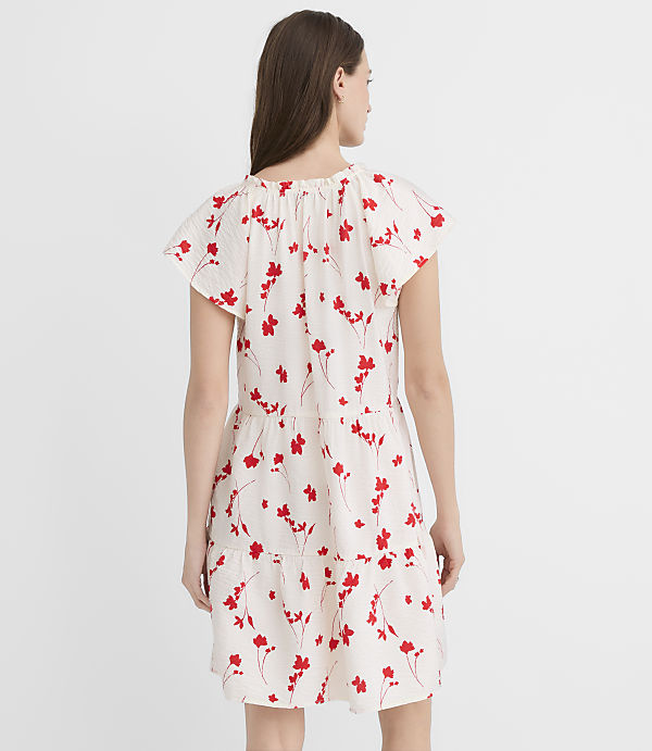 A woman in a white dress with red floral print stands against a plain white background.