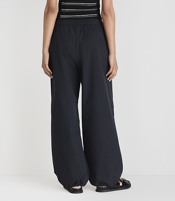 A person wearing dark, wide-leg pants and sandals stands against a plain white background.