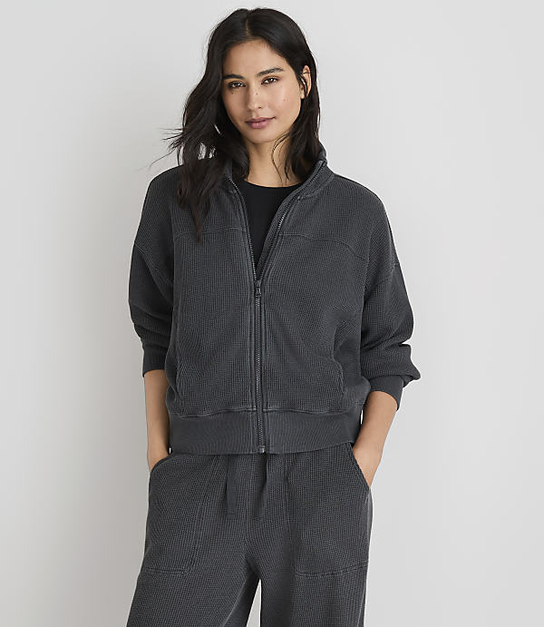 A woman in a dark grey zip-up jacket and matching pants stands against a plain white background.