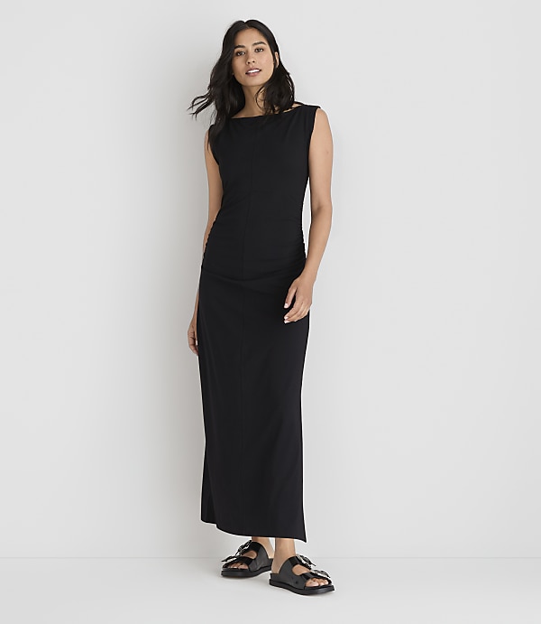 A woman in a black maxi dress and sandals stands against a plain white background.