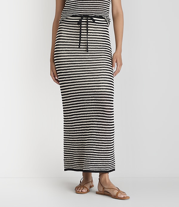 A person stands wearing a black and white striped maxi skirt and sandals against a plain white background.