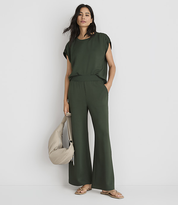 A woman in a green top and wide-leg pants stands against a plain white background, holding a beige bag.