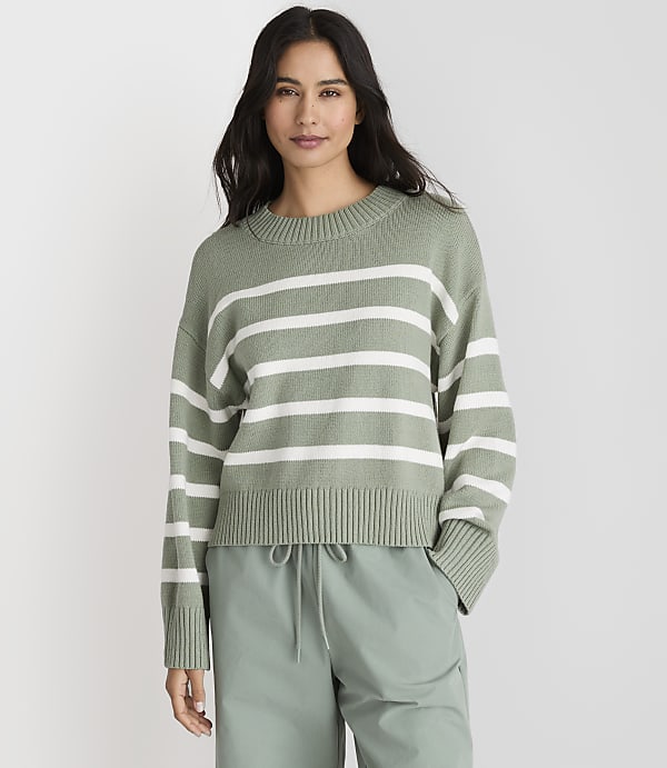 A woman in a green and white striped sweater and light green pants stands against a plain gray background.