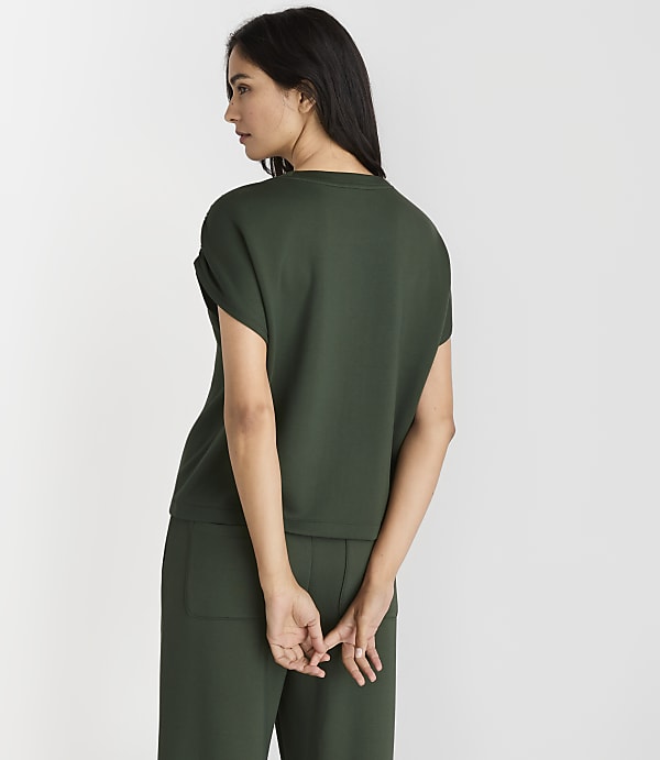 A woman in a dark green top and pants stands with her back to the viewer against a plain white background.