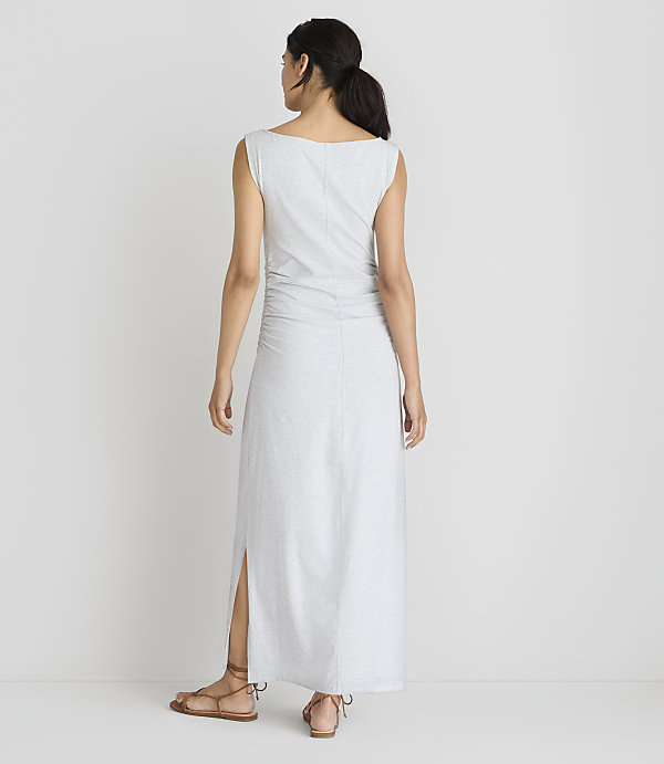 A woman in a light gray maxi dress with ruched detailing and a side slit stands with her back to the camera against a plain white background.