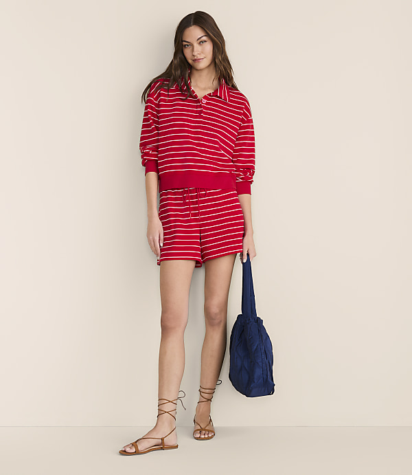 A woman in a red and white striped polo shirt and shorts stands against a plain, light-colored background, holding a blue bag.