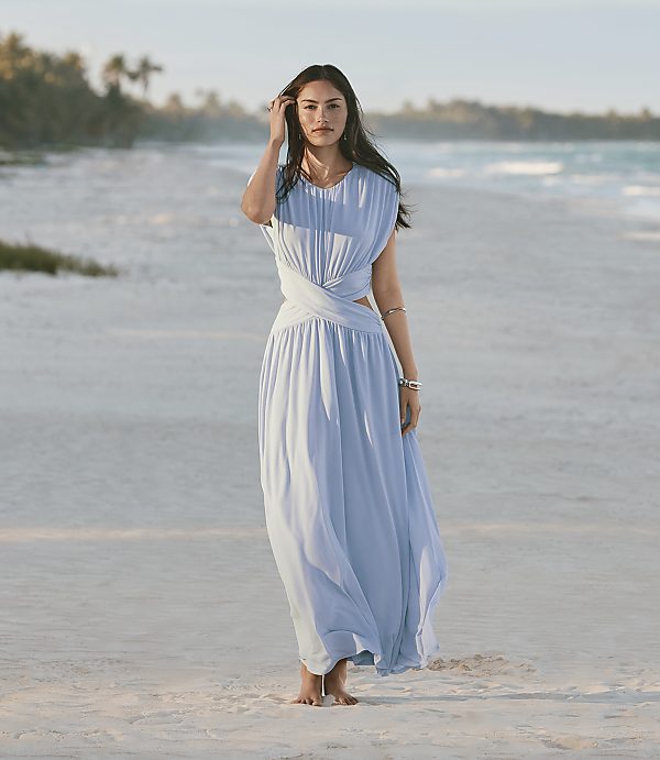 A woman in a light blue dress stands on a sandy beach with the ocean and palm trees in the background.