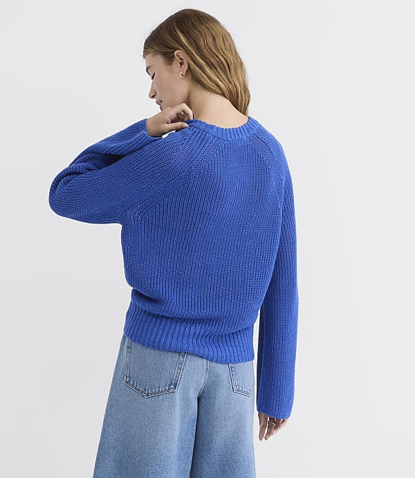 A woman in a blue sweater and wide-leg jeans stands against a plain white background.