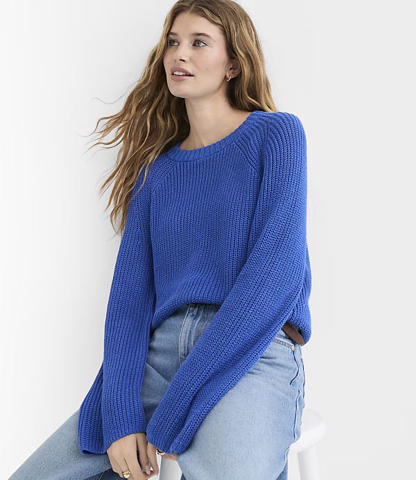 A young woman in a blue sweater and jeans sits against a plain white background.