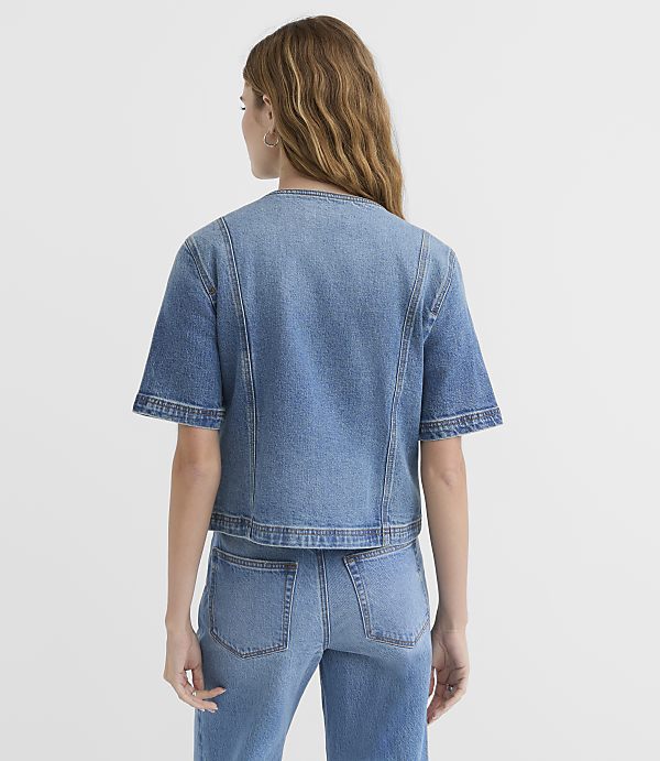 A woman in a denim jacket and jeans stands against a plain white background.