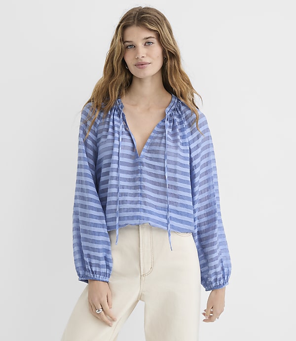 A woman in a blue striped blouse and cream pants stands against a plain white background.
