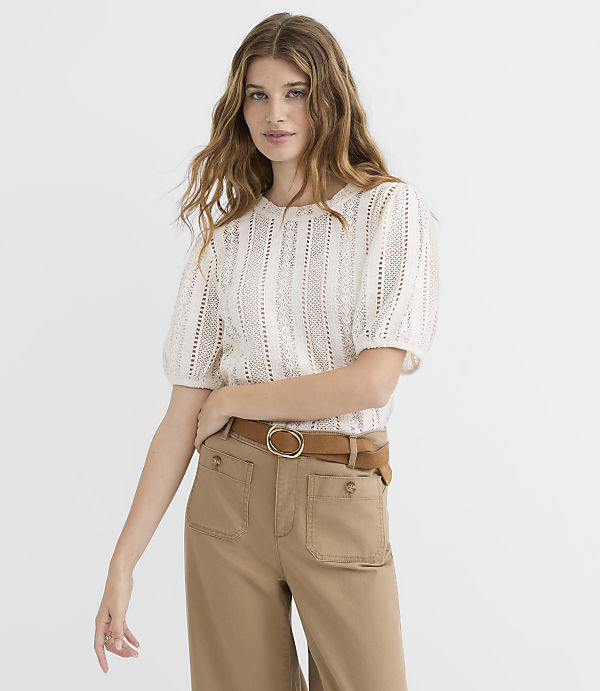 A woman with long, wavy blonde hair wears a cream-colored, open-knit top and a brown crossbody bag, standing against a plain white background.