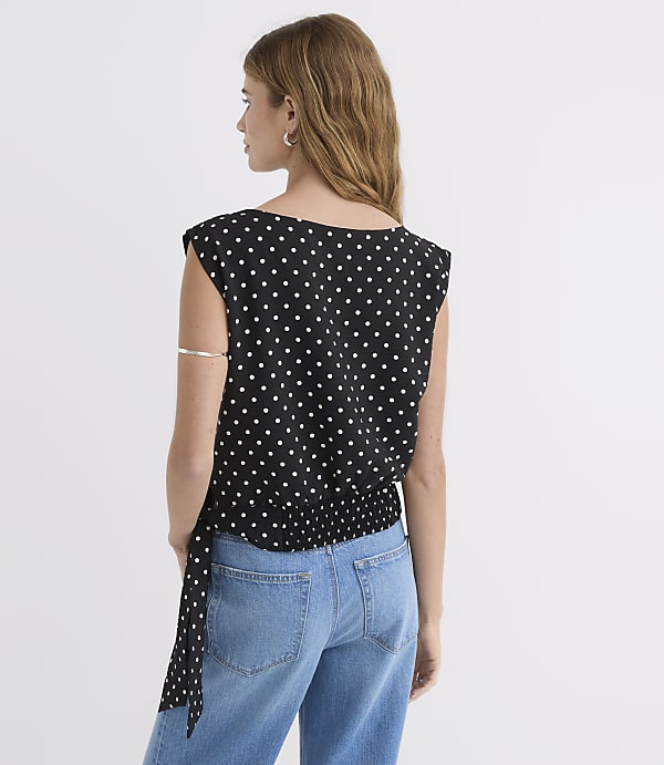 A woman in a black polka dot top and blue jeans stands against a plain white background.