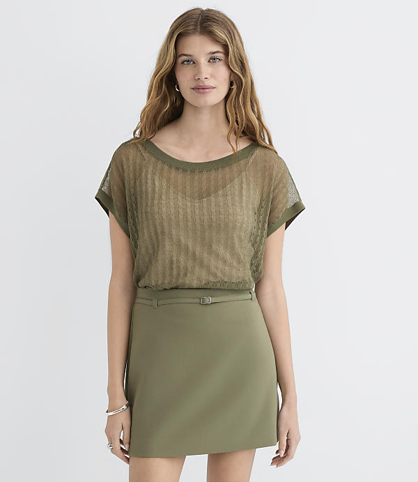 A woman in a sheer olive green top and olive green skirt stands against a plain white background.