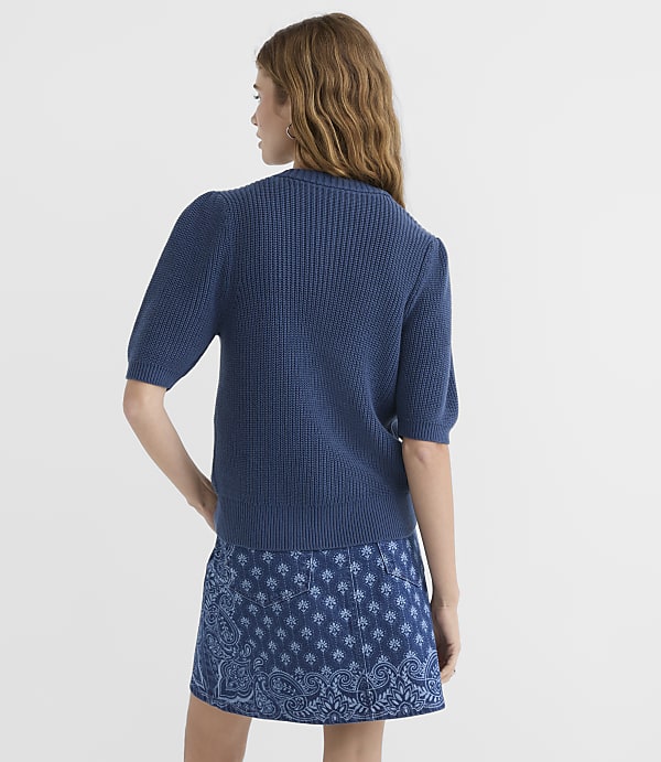 A woman in a blue ribbed sweater and a patterned blue skirt stands against a plain white background.