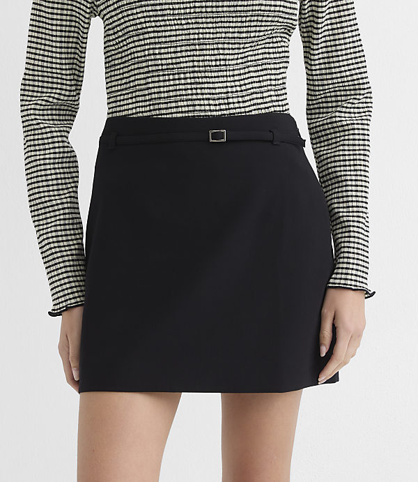 A black mini skirt with a belted waist is worn by a person with a striped long-sleeved shirt against a plain gray background.