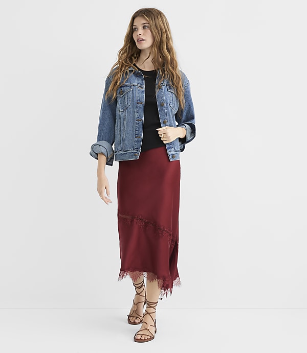 A woman in a denim jacket and maroon skirt stands against a plain white background.
