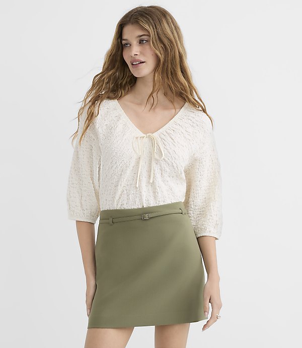 A young woman in a textured cream top and olive green skirt stands against a plain white background.