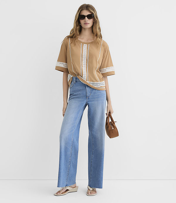 A woman in a tan top and wide-leg jeans stands against a plain white background, holding a small brown purse.
