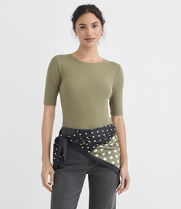 A woman in a green ribbed top and dark jeans stands against a plain white background, with a polka-dotted scarf tied around her waist.